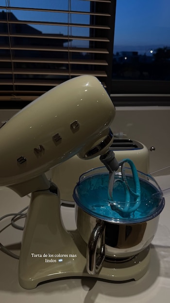 Batidora de pie Smeg color crema mezclando sustancia azul brillante en un tazón. Al fondo, una ventana con persianas y cielo oscuro al atardecer