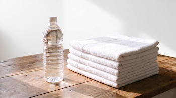 Una botella de plástico con agua transparente está de pie junto a una pila de seis toallas blancas dobladas sobre una mesa de madera. Fondo blanco.