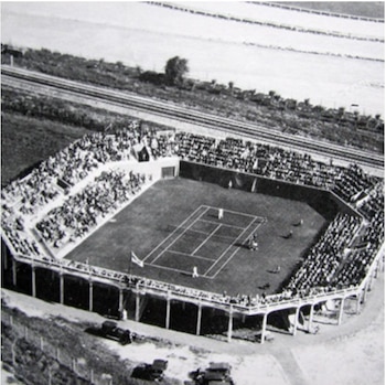 Vista aérea en blanco y negro de una cancha de tenis con personas en las tribunas y automóviles estacionados en el exterior