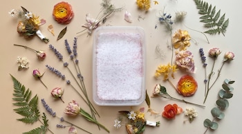 Vista superior de varias flores, ramas de lavanda y helechos distribuidos alrededor de un recipiente rectangular con polvo blanco.