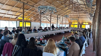 Vista interior de un salón rústico con techo de madera, donde un grupo grande de personas se sienta en largas mesas para un evento, con un orador en el centro