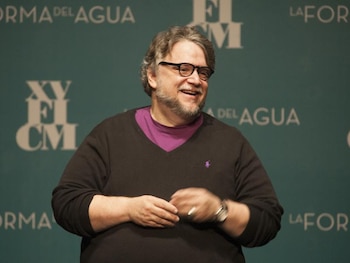 Guillermo del Toro sonríe vistiendo anteojos y un suéter oscuro frente a un fondo verde con el texto La forma del agua y XV FICM