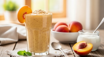 Primer plano de un licuado de durazno, avena y yogur en un vaso, con una rebanada de durazno y avena encima. Hay duraznos, yogur y menta sobre una mesa de madera.