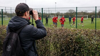 Vista trasera de una persona con gorra y mochila, oculta parcialmente tras un seto, observando un entrenamiento de fútbol con binoculares a través de una valla.
