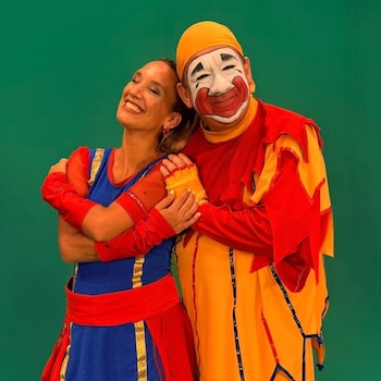 Una mujer en traje azul y rojo abraza a un hombre disfrazado de payaso con maquillaje blanco y rojo, vistiendo un traje amarillo y rojo, sobre fondo verde