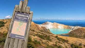 Un cartel de madera con mapa en japonés en primer plano frente a un terreno montañoso y un lago de color turquesa dentro de un cráter