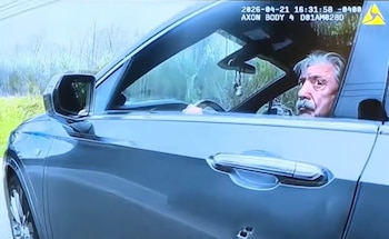Un hombre canoso con bigote mira a la cámara desde la ventanilla del conductor de un automóvil gris que presenta marcas de disparos