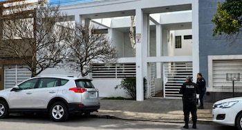Dos agentes de policía uniformados frente a una casa moderna de color blanco y gris con dos autos plateados estacionados en la calle