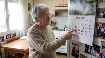 Adulta mayor con gafas y cabello gris señalando el día 18, circulado en rojo, de un calendario de Mayo de 2026 en la puerta de un refrigerador.
