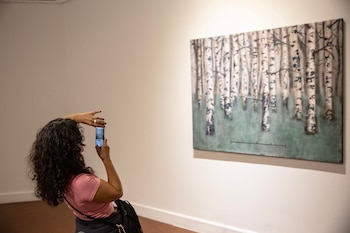 Vista trasera de una persona con cabello rizado y camiseta rosa fotografiando una pintura de un bosque de abedules en una pared blanca de galería