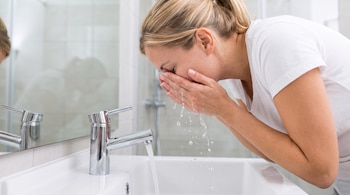Una mujer con camiseta blanca se lleva agua a la cara con las manos sobre un lavabo blanco con grifo cromado.
