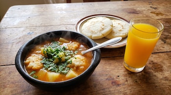 Sopa de arepa humeante en plato negro con cilantro, dos arepas blancas en plato marrón y un vaso de jugo amarillo sobre una mesa de madera.