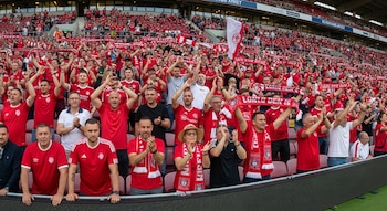 Panorámica de una hinchada mediana de fútbol, mayormente hombres, vestidos con camisetas rojas, ondeando bufandas y aplaudiendo en un estadio.