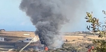 Helicóptero volando entre una gran columna de humo negro por un incendio en un terreno árido con pequeñas casas blancas. Se observa un chorro de agua
