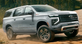 Vehículo todoterreno Chevrolet Suburban gris claro circulando en un camino de tierra. Fondo con árboles y vegetación