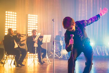Un cantante inclinado sostiene un micrófono en un escenario iluminado con varios músicos al fondo