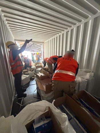 Varios trabajadores con chalecos de seguridad y cascos dentro de un contenedor de envío blanco. Uno apunta hacia el techo, mientras otros manejan cajas y bultos