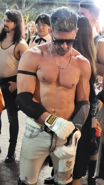 Hombre musculoso sin camiseta con gafas de sol, cadena plateada, guantes de lucha y jeans claros posa en un evento al aire libre