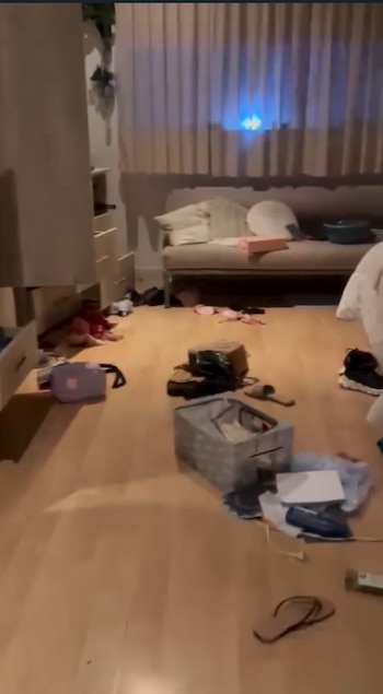 Piso de madera clara con ropa, bolsos y objetos esparcidos. Mueble de cajones abiertos, banco o cama, y cortinas en el fondo