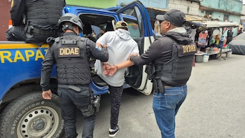 Tres agentes de policía, dos de ellos con chalecos que dicen 'DIDAE', esposan a un hombre y lo suben a la parte trasera de una camioneta azul