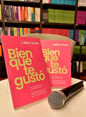 Dos libros de tapa rosa intenso con el título "Bien que te gustó" y el nombre "Julieta Saulo" junto a un micrófono, con estanterías llenas de libros de colores al fondo