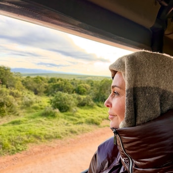Mujer de perfil mirando por la ventana de un vehículo. Viste chaqueta marrón brillante y capucha tejida. Exterior con cielo nublado, vegetación y camino de tierra