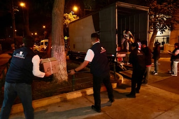 Vista nocturna de varios hombres con chalecos de la ONPE descargando o cargando cajas de un camión grande. Hay árboles y luces de la calle al fondo
