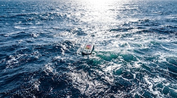 Superficie del océano con olas y reflejos de luz solar, un termómetro digital flotante en el centro de la imagen que muestra una pantalla con colores cálidos.
