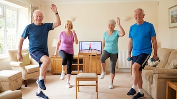 Cuatro adultos de 60 años sonríen mientras hacen ejercicio en su sala, imitando una rutina de aeróbicos en la TV. Dos hombres y dos mujeres en ropa deportiva.