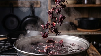 Primer plano de flores de hibisco secas de color rojo oscuro cayendo en una olla de agua hirviendo con vapor. Fondo de cocina oscuro y borroso con sartenes.