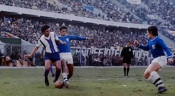 Tres futbolistas disputan el balón en una cancha de césped frente a un árbitro y tribunas llenas de espectadores en un estadio