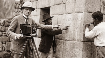Hombre con sombrero de explorador y cámara en trípode observa a dos hombres, uno señala el texto "A. LIZÁRRAGA 1902" en una pared, otro lo borra con cincel.