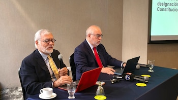 Dos hombres con traje y corbata están sentados en una mesa con micrófonos, laptops y vasos. El hombre de la izquierda sostiene un micrófono y habla