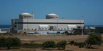 Vista general de la Estación Nuclear de Koeberg con dos cúpulas blancas y un gran edificio gris industrial al lado del mar. Vegetación seca en primer plano