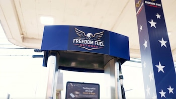 Un dispensador de combustible azul y plata de una estación Freedom Fuel Network con logo de águila; un pilar azul con estrellas blancas y una pantalla con texto