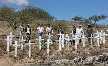 Grupo de personas, mayormente indígenas, de pie en un terreno árido entre una multitud de cruces blancas con nombres, bajo un cielo azul despejado, con árboles secos al fondo
