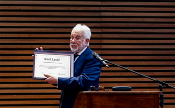 Raúl Lavié sostiene un diploma enmarcado detrás de un atril con micrófono frente a una pared de listones de madera