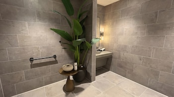 Un baño interior muestra azulejos rectangulares grises en las paredes y cuadrados en el suelo, una planta grande, una mesa auxiliar pequeña y un lavamanos con espejo iluminado.