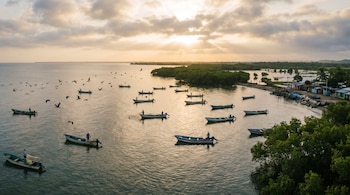 Vista aérea de la bahía de Jiquilisco al amanecer con sol brillante. Múltiples botes de pesca y pescadores en aguas tranquilas, con aves en vuelo y manglares.