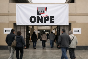 Vista frontal de la entrada de un local de votación con un gran cartel de la ONPE. Varias personas desenfocadas ingresan al edificio beige, con flechas direccionales en las paredes.