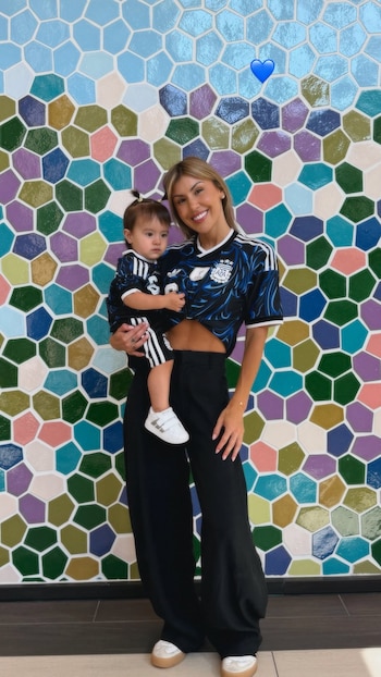 Mujer rubia sonriente sostiene un bebé. Ambos visten camiseta de la Selección Argentina negra. Fondo: pared con mosaicos de colores y un corazón azul