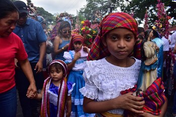 Primer plano de una niña con tocado colorido y blusa blanca, sosteniendo una estatua de la Virgen María, en medio de una multitud durante una procesión con flores y plantas artificiales