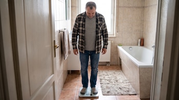 Hombre sobre balanza digital en baño con azulejos, bañera y ventana. Viste camisa a cuadros, camiseta, pantalones y calcetines.