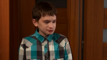Primer plano de un niño de cabello castaño corto que viste una camisa a cuadros de colores turquesa, gris, blanco y negro, sobre un fondo de madera marrón