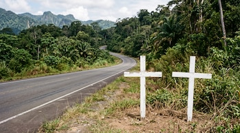 Dos cruces blancas de madera en el borde de una carretera asfaltada rodeada de vegetación y montañas al fondo.