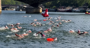 Numerosos nadadores con gorros blancos nadan en un río con boyas rojas y naranjas, y un puente de piedra al fondo con embarcaciones