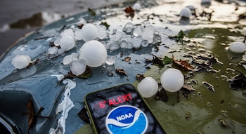 Bolas de granizo de hasta 7,5 cm sobre el capó abollado de un auto gris metálico, rodeadas de agua y hojas, junto a un móvil con el logo NOAA y una alerta roja.