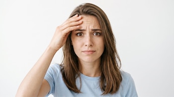 Primer plano de mujer joven de pelo castaño, camiseta azul, con expresión de confusión y mano derecha en la frente, sobre fondo blanco liso