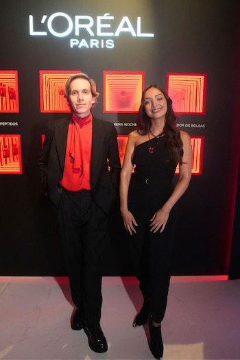 Hombre con traje negro y camisa roja, mujer con mono negro y collar rojo posan frente a pared negra con logo L'Oréal y recuadros rojos