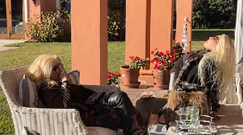 Dos mujeres de cabello rubio, sentadas en sillas de mimbre en un patio exterior, con columnas color salmón, plantas y césped verde.